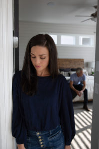 woman in foreground looks down sadly as husband sits on bed