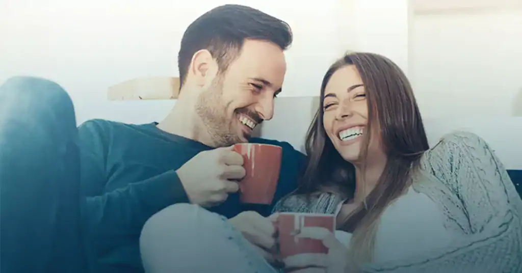 Happy couple laughing together on couch holding coffee mugs