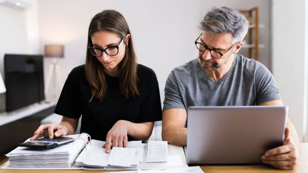 A woman with glasses uses a calculator while reviewing paperwork at a table, next to a man looking at a laptop.